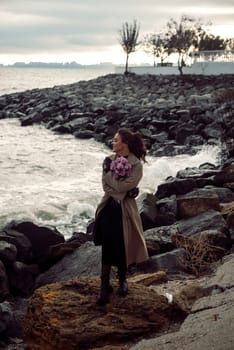 young girl on the seashore around the rocks. she is dressed in a beige trench coat, she is dreaming on the seashore among the flowers of the chrysanthemum variety. a chair decorated with blue fabric
