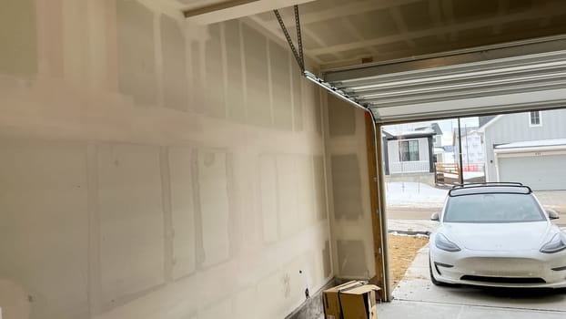 Castle Pines, Colorado USA - January 18, 2023-Interior of a garage in a single-family home under construction, showcasing unfinished drywall and an open garage door. A white electric car is visible in the driveway, with neighboring homes in a suburban setting.