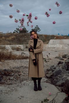 young girl on the seashore around the rocks. she is dressed in a beige trench coat, she is dreaming on the seashore among the flowers of the chrysanthemum variety. a chair decorated with blue fabric