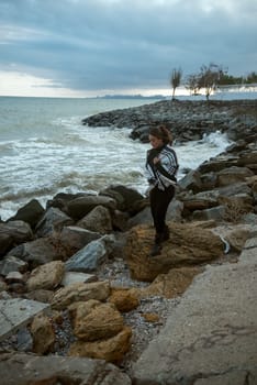 young girl on the seashore around the rocks. she is dressed in a beige trench coat, she is dreaming on the seashore among the flowers of the chrysanthemum variety. a chair decorated with blue fabric