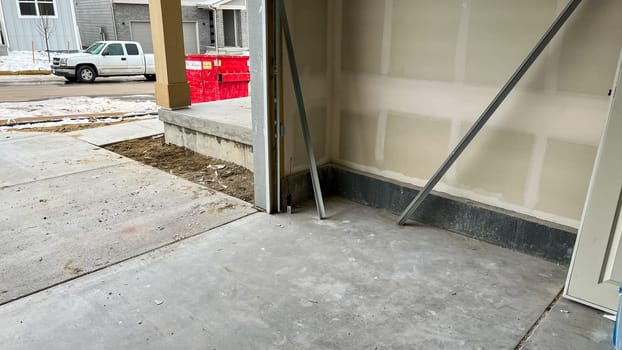 Castle Pines, Colorado USA - January 18, 2023-Interior view of a garage under construction in a new single-family home. The space features unfinished drywall, concrete flooring, and construction materials leaning against the wall. The open garage door reveals a driveway, suburban neighborhood, and a red construction dumpster.