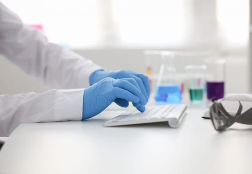 Employee chemical laboratory teacher chemist knocks his fingers keyboard makes notes electronic journal records. Analysis data from reactions examining test tubes with the substance arm in gloves.