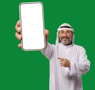 Arab man smiles and showcases mobile phone with blank white display, offering copy space for app advertising, services and information. Happy man isolated against green background. . High quality photo