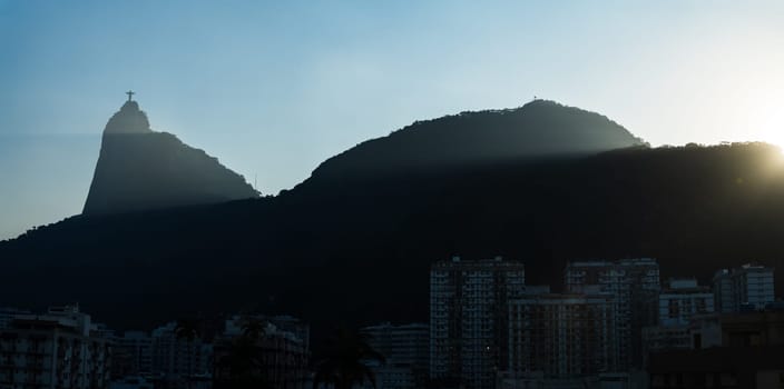 Stunning sunset view of Rio de Janeiro's mountains and Christ the Redeemer, with sunrays and silhouettes offering ample text space.
