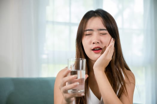 Beautiful female toothache and dental problems touching cheek feeling pain after drinking cold water, Asian young woman with sensitive teeth holding glass of cold water at home in living room, pain