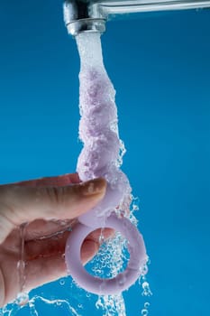 Woman holding lilac anal beads under running water on blue background. Sex toy hygiene concept