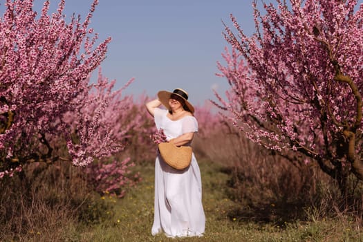 Woman blooming peach orchard. Against the backdrop of a picturesque peach orchard, a woman in a long white dress and hat enjoys a peaceful walk in the park, surrounded by the beauty of nature