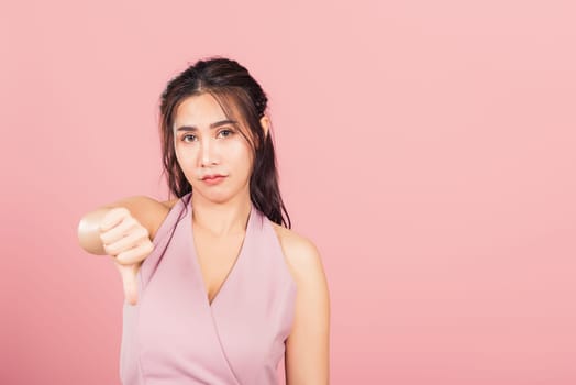 Portrait Asian beautiful young woman unhappy, negative gesture showing finger thumbs down or dislike sign, studio shot isolated on pink background, Thai female rejection unlike with copy space