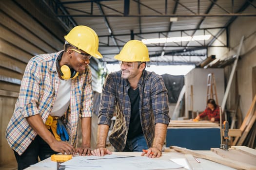 Two carpenter man wear helmet meeting planning job together at carpentry workshop, craftsmen talking checklist and plan on blueprint paper in woodshop, teamwork, National Carpenters Day