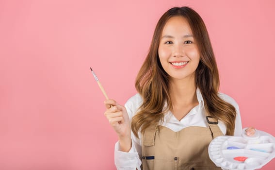 Asian beautiful young woman artist holding brush and paint palette, Happy female painting using paintbrush and palette with colors, studio shot isolated on pink background, Paintings and art equipment
