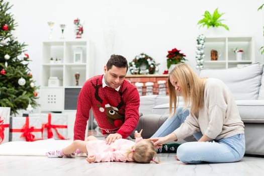 A man and a woman sit on the floor and play with their little daughter. A little girl in a pink dress lies on the floor. The room is decorated with Christmas decorations and a Christmas tree.