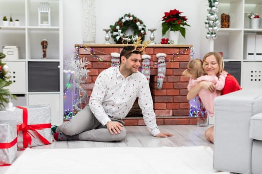 A loving mother hugs her little daughter while sitting by the fireplace. The whole family is spending time together in a festive mood. Dad sits on the floor by the fireplace. The family of three is smiling.