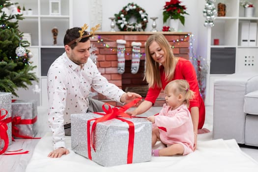 A woman, a man and a little girl sit in a room full of Christmas decorations. The whole family is sitting around a large gift. The box is wrapped with a red ribbon. The little girl pulls at the ribbon.