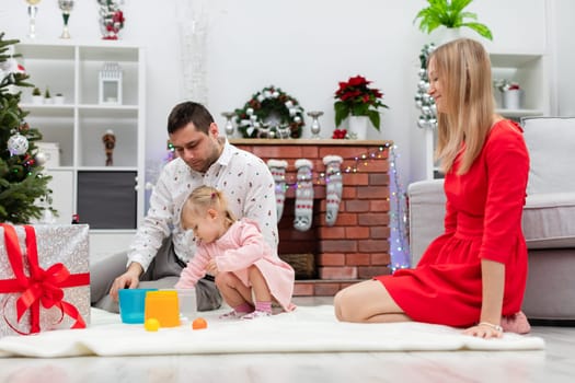 A blonde girl in a red dress kneels on a white carpet. Mom, dad and little daughter are playing by the Christmas tree. Under the Christmas tree lie gifts. The room is decorated with Christmas ornaments.