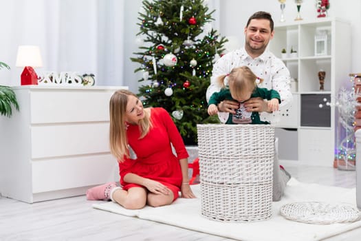 The room is decorated with Christmas ornaments. A dad lifts up his little daughter. A man takes the baby out of a wicker basket. A woman in a red dress sits on the floor and laughs with her mouth open.