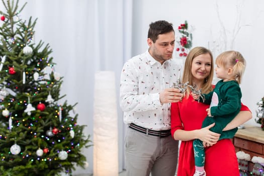 A woman in a red dress holds a little girl in her arms. A man dressed in a patterned shirt stands behind the woman. The family is spending time in a festive atmosphere. A dressed Christmas tree can be seen in the background.