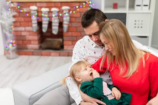 A man, a woman and a little girl sit together on a gray couch. The woman holds her lying daughter in her lap. The blurry background shows a brick fireplace decorated with Christmas lights.