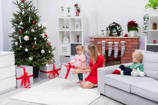 A daughter and son are staying with their mother in a room decorated with Christmas ornaments. The little boy is sitting on the couch. The little girl is standing by the Christmas tree, and her mother is handing her a gift wrapped with a bow.
