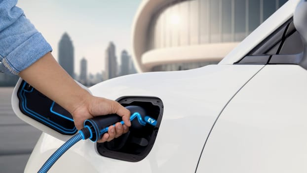 Asian man insert EV charger and recharge his electric car from future charging station with blurred background of cityscape. Smart and futuristic sustainable energy infrastructure. Peruse