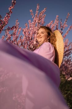 Woman blooming peach orchard. Against the backdrop of a picturesque peach orchard, a woman in a long pink dress and hat enjoys a peaceful walk in the park, surrounded by the beauty of nature