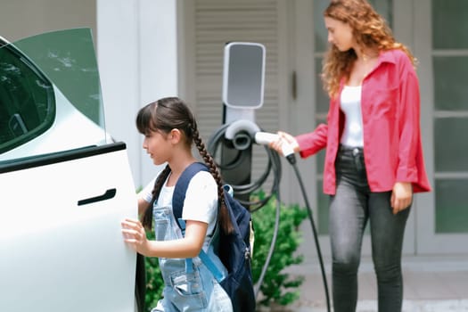 Happy little young girl learn about eco-friendly and energy sustainability as she help her mother recharge electric vehicle from home EV charging station. EV car and modern family concept. Synchronos