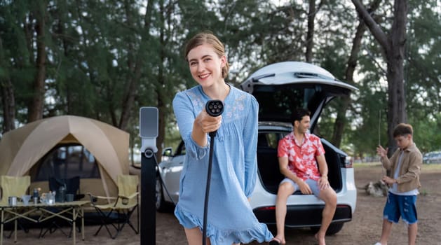 Outdoor adventure and family vacation camping at sea travel by eco friendly car. Cheerful woman or mother holding, pointing EV charger point with playful and happiness posture in campsite. Perpetual