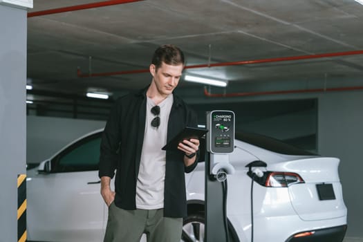 Young man travel with EV electric car to shopping center parking lot charging in downtown city showing urban sustainability lifestyle by green clean rechargeable energy of electric vehicle innards