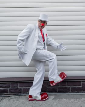 An elderly man in a white suit, huge boots and a clown nose walks funny