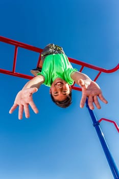 A happy young boy hangs upside down from the monkey bars.