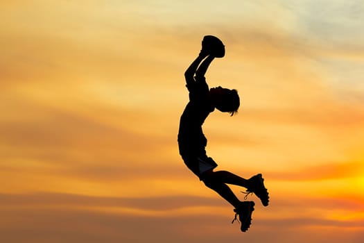 A young boy in silhouette is playing football at sunset against an orange sky.