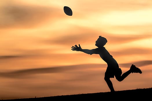 A young boy in silhouette is playing football at sunset against an orange sky.