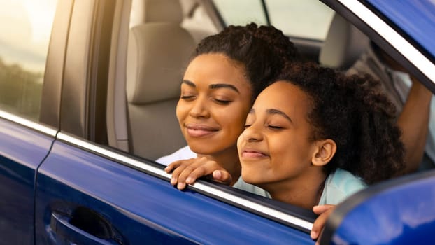 Joyful Black Mother And Daughter Sitting In Automobile, Enjoying Road Trip Smelling Fresh Air In Opened Window, Family Having Summer Adventure Traveling By Car. Transportation Concept