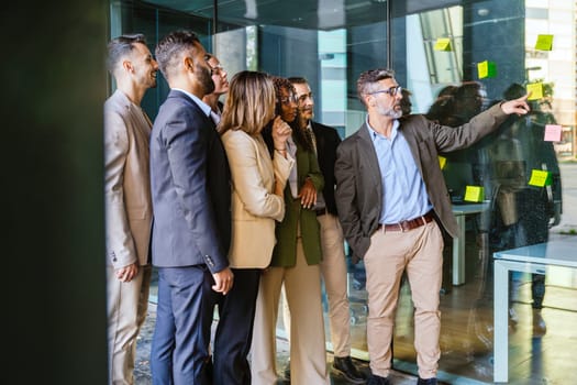 A diverse group of business professionals brainstorming in a modern office. A man points to sticky notes on a glass wall as the team collaborates with focus and engagement.