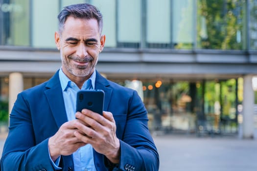Smiling middle-aged businessman in a blue suit holding and using a smartphone outdoors in a modern urban setting.