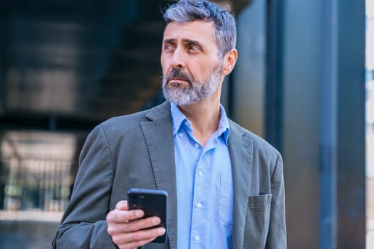 Middle-aged businessman in smart casual attire holding a smartphone, looking away thoughtfully in a modern urban environment.
