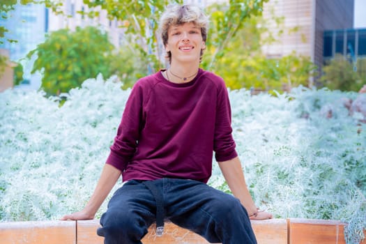 Young man in a burgundy shirt sitting on a bench, smiling in an urban park setting surrounded by greenery and plants.