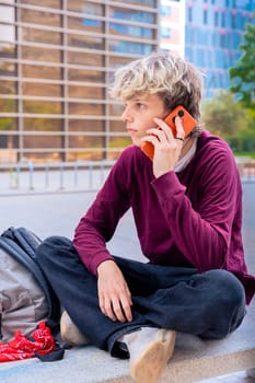 A young man with blond hair sits casually outdoors, talking on his smartphone. Urban background with modern buildings, a backpack, and a relaxed vibe, symbolizing connectivity.