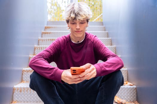 Young man in a burgundy shirt sitting on metal stairs, focused on his smartphone, with autumn leaves scattered around in an outdoor urban setting.