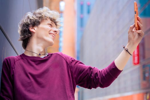 A cheerful young man with blond hair takes a selfie using his smartphone in a modern urban setting. Casual and vibrant scene highlighting youth and digital connectivity.