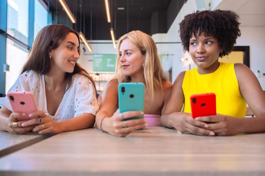 A group of three diverse women sitting at a table in a modern cafe, holding smartphones and smiling. Casual and cheerful atmosphere representing friendship and digital connectivity.