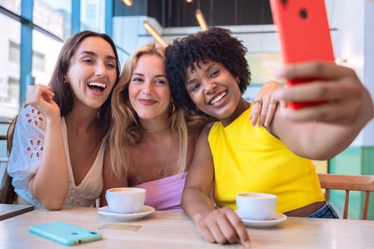 Three diverse women smiling and taking a selfie together while enjoying coffee in a trendy cafe. Cheerful and vibrant scene representing friendship, happiness, and modern lifestyle.