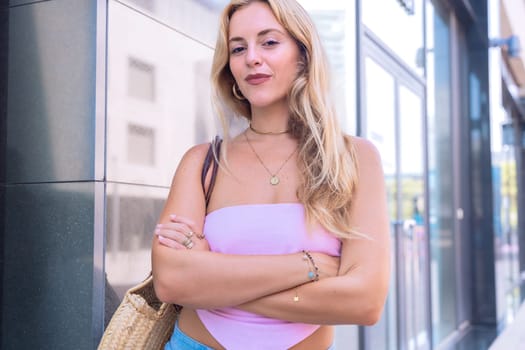 A blonde woman standing confidently with arms crossed, wearing a trendy summer outfit. Captured in an urban setting with a casual and fashionable vibe, showcasing modern elegance.