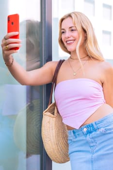 A cheerful blonde woman wearing a pink top and jeans takes a selfie with her smartphone. Captured in an urban setting, showcasing casual style, confidence, and modern connectivity.