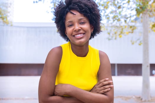 A cheerful young woman with natural curly hair wearing a yellow top smiles confidently while posing outdoors. Bright and vibrant setting, showcasing joy, beauty, and individuality.