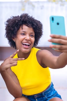 A joyful young woman with curly hair wearing a yellow top takes a selfie outdoors, smiling and gesturing peace. Vibrant scene showcasing happiness, individuality, and modern lifestyle.Vertical