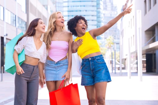 A group of three cheerful women carrying shopping bags, pointing and laughing while strolling in a modern urban area. Vibrant scene showcasing friendship, shopping, and fun moments.