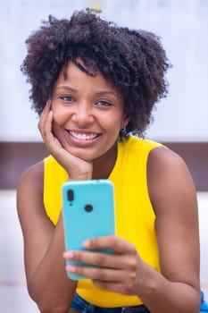 A young woman with natural curly hair wearing a yellow top rests her face on her hand while holding a smartphone. Calm and reflective moment captured in a bright outdoor setting.