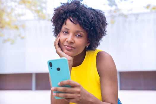 A young woman with natural curly hair wearing a yellow top rests her face on her hand while holding a smartphone. Calm and reflective moment captured in a bright outdoor setting.