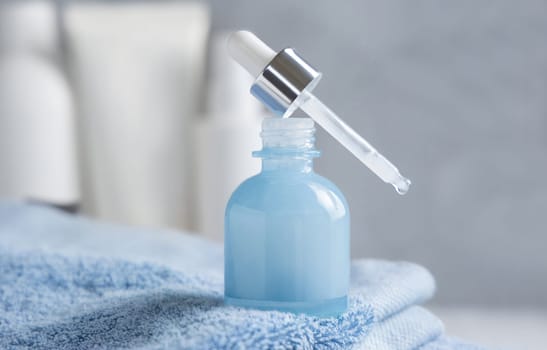 Drop falling from pipette set on opened blue glass dropper bottle on blue towel against cosmetic bottles and grey wall close up, mockup. Natural serum or essence
