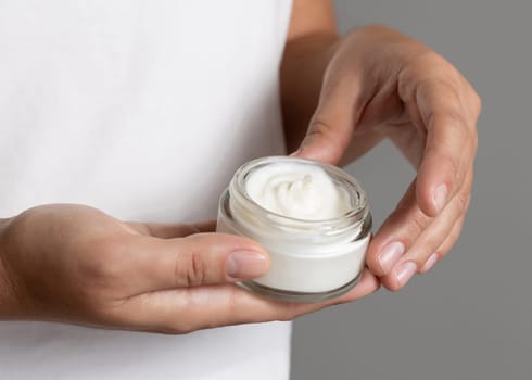 Female hand holding cream jar against white t-shirt close up. Natural product for facial care, everyday skincare routine. Opened cosmetic Container. Spa and Wellness concept